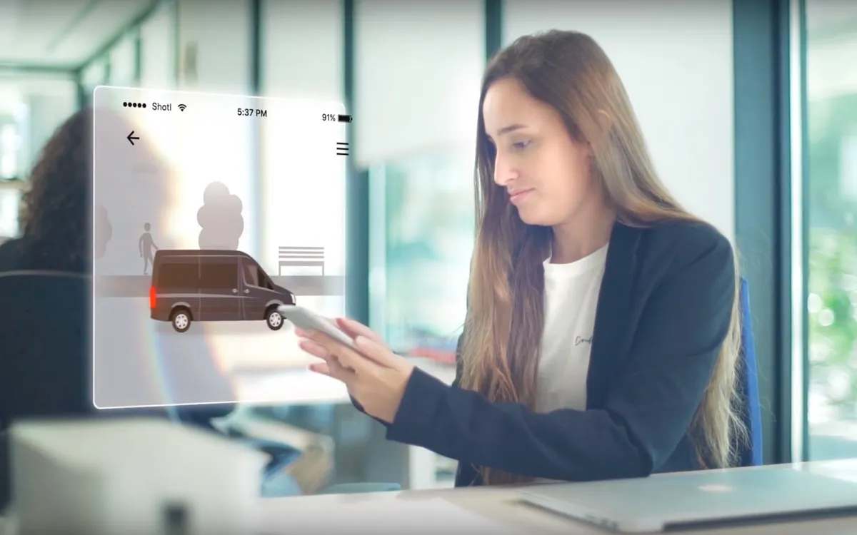 Girl holding a phone displaying a van on screen, in an office setting.
