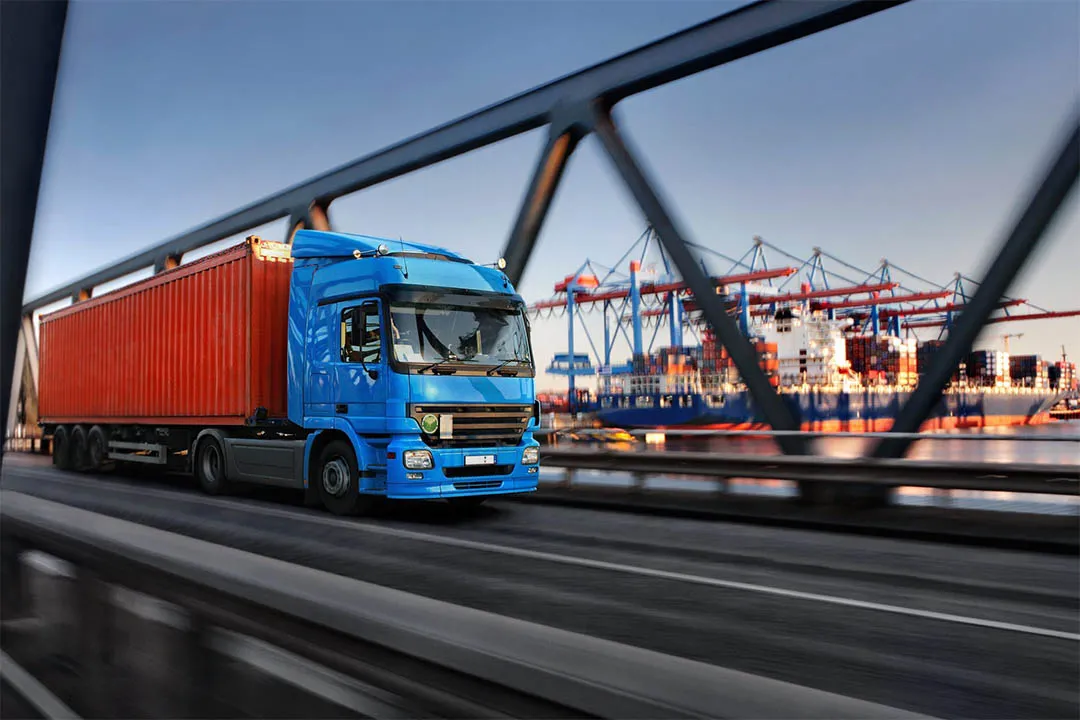 Blue semi-truck hauling a red container on a highway with industrial cranes in the background.