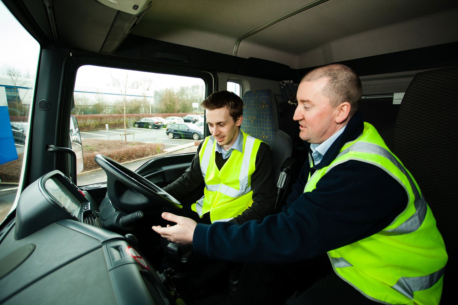 Two people wearing high-visibility vests inside a vehicle, one in the driver's seat.